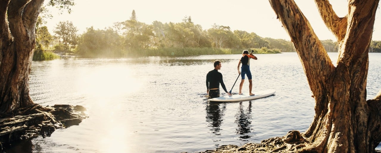 Dieťa stojí na paddle boarde a padluje po jazere, zatiaľ čo ho otec, ktorý stojí vo vode pridržiava.